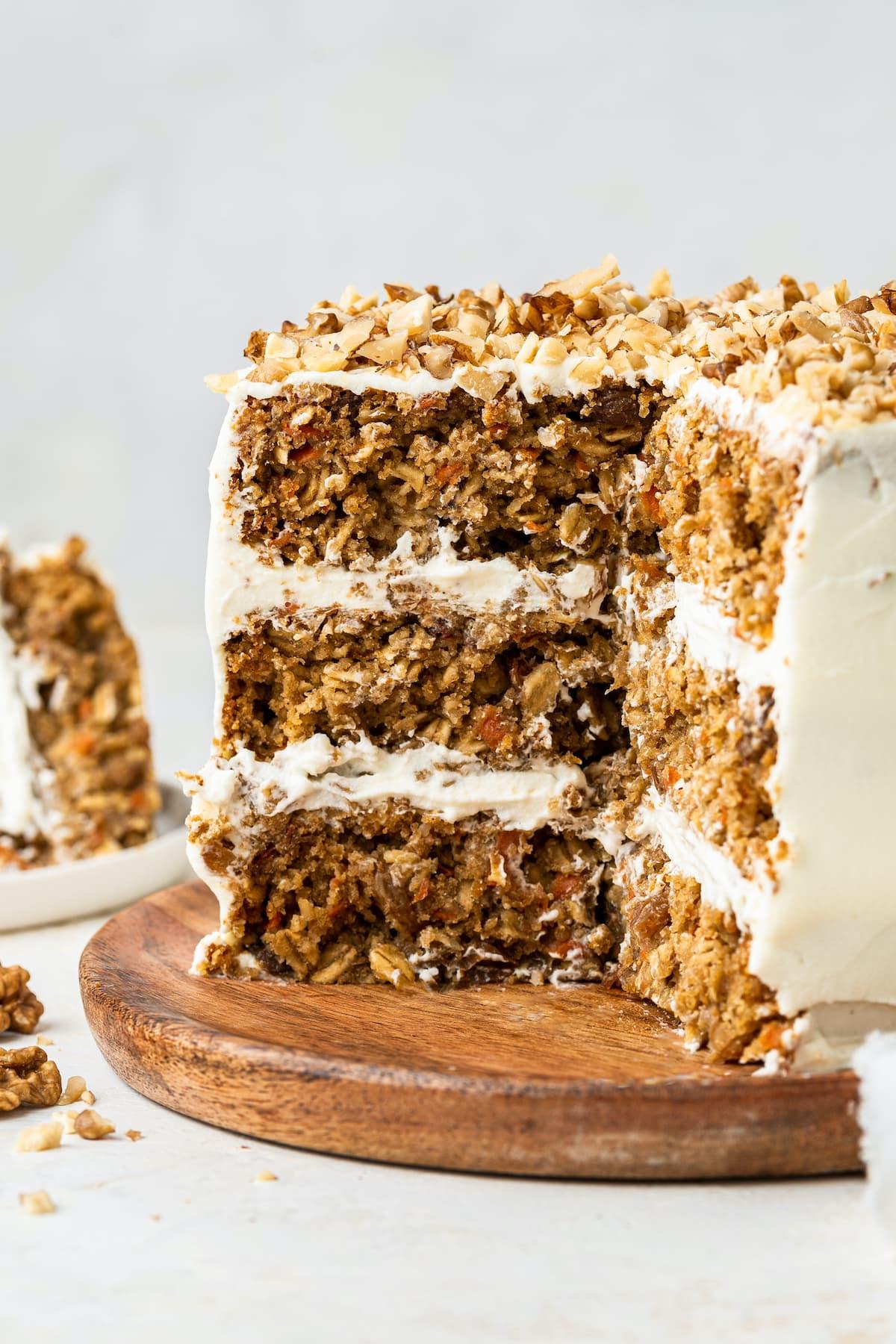 Κέικ βρώμης και καρότου 2 3-layer oatmeal carrot cake, frosted with chopped walnuts on top served on a wooden board. A few slices of the cake have been cut out exposing the inside of the cake.