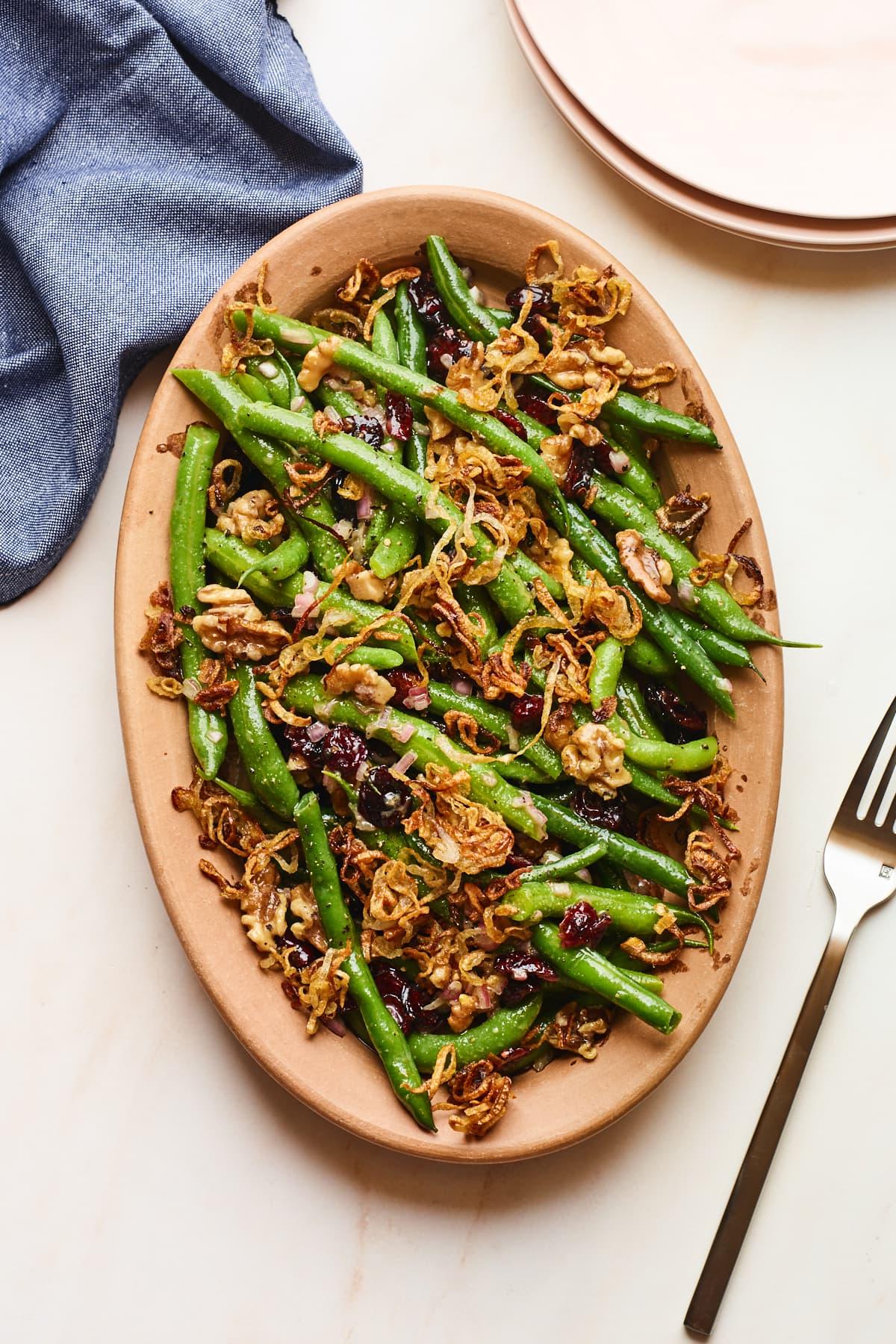Φασολάκια Σαλάτα 2 An overhead photo looking down onto a serving dish containing green bean salad topped with fried shallots. A fork is resting next to the serving dish, as well as a blue linen dishcloth.