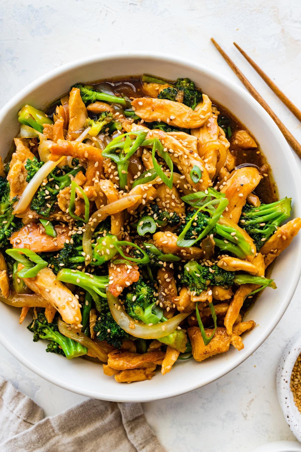 **Κοτόπουλο και Μπρόκολο Σοτέ** 2 Κοτόπουλο stir fry με μπρόκολο σε λευκό μπολ, γαρνιρισμένο με σουσάμι και φρέσκο κρεμμυδάκι, με ξύλινα chopsticks δίπλα.