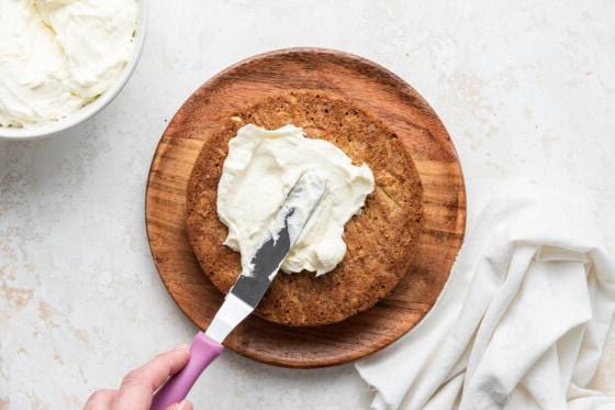 Woman's hand frosting one layer of the oatmeal carrot cake with a small cake spatula with a pink handle.