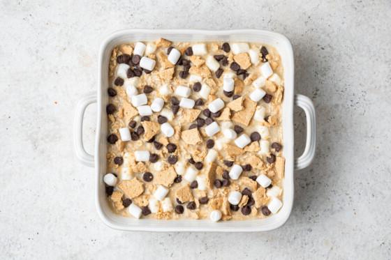 Ένα λευκό τετράγωνο ταψί με την ψητή βρώμη S'mores μέσα πριν το ψήσιμο στο φούρνο. Στολισμένο με κομματάκια σοκολάτας, μίνι marshmallows και τριμμένα μπισκότα graham.