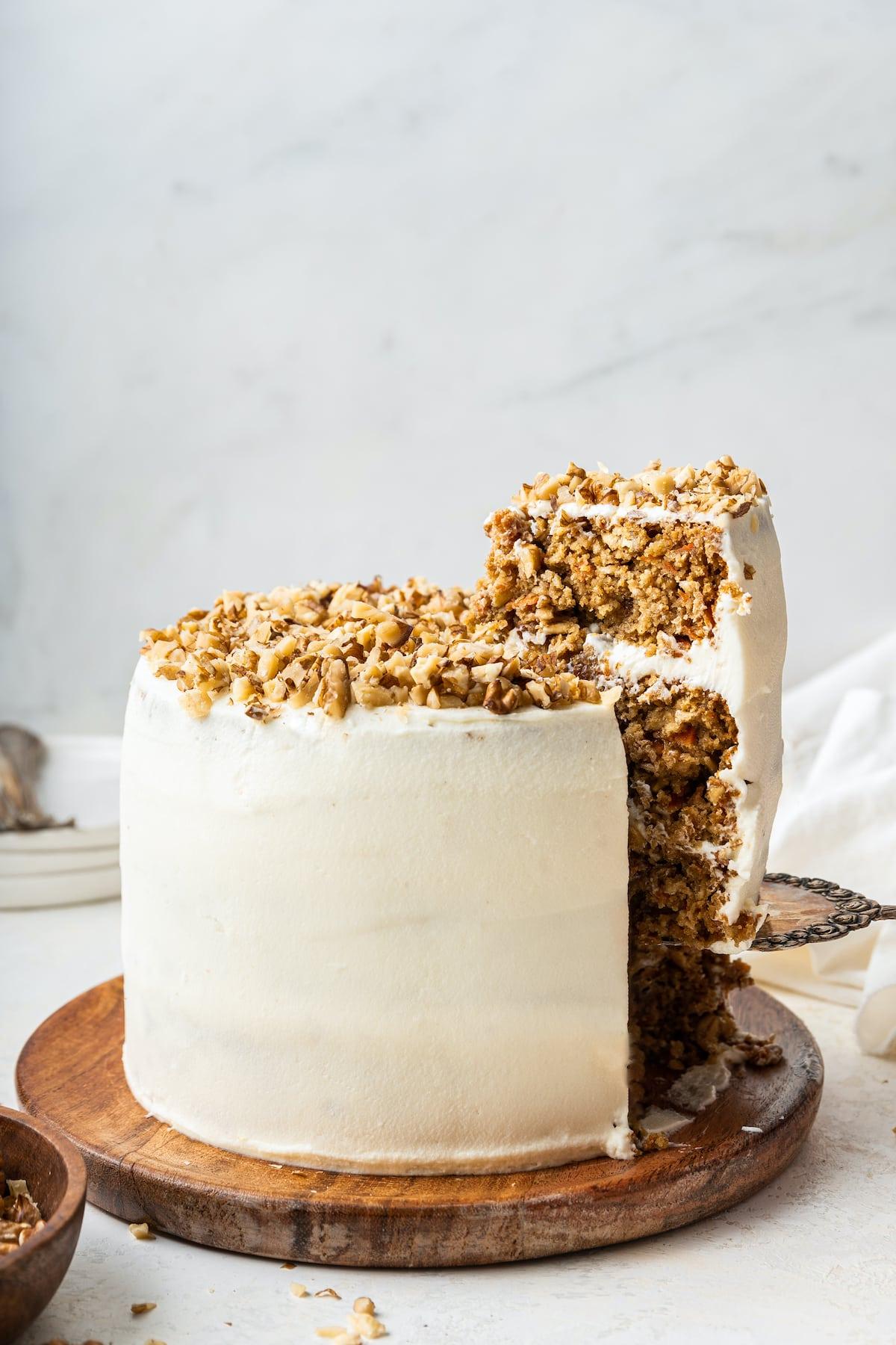 Κέικ βρώμης και καρότου 5 Oatmeal carrot cake on a wooden board with a slice being removed from it. The cake is frosted in a cream cheese frosting and has chopped walnuts on top.
