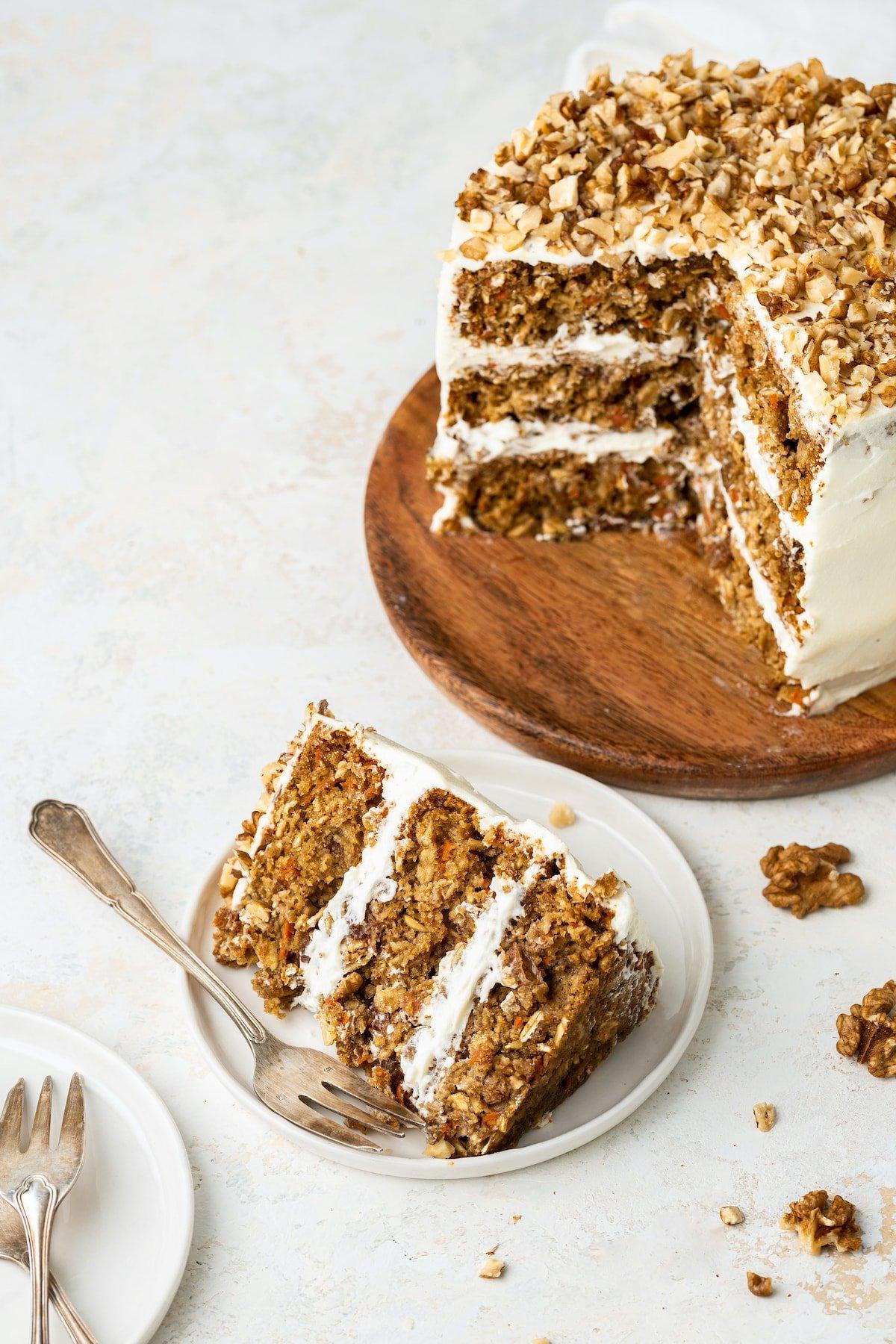 Κέικ βρώμης και καρότου 6 A slice of a 3-layer oatmeal carrot cake served on a white plate with a fork. The full 3-layer oatmeal carrot cake is behind the plate on a wooden board with the center exposed.