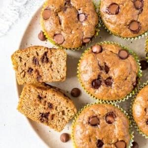 Μάφινς κολοκυθιού χωρίς αλεύρι 6 Πλάκα με muffins κολοκυθιού χωρίς αλεύρι και σταγόνες σοκολάτας. Ένα muffin είναι κομμένο στη μέση και φαίνονται οι σταγόνες σοκολάτας.