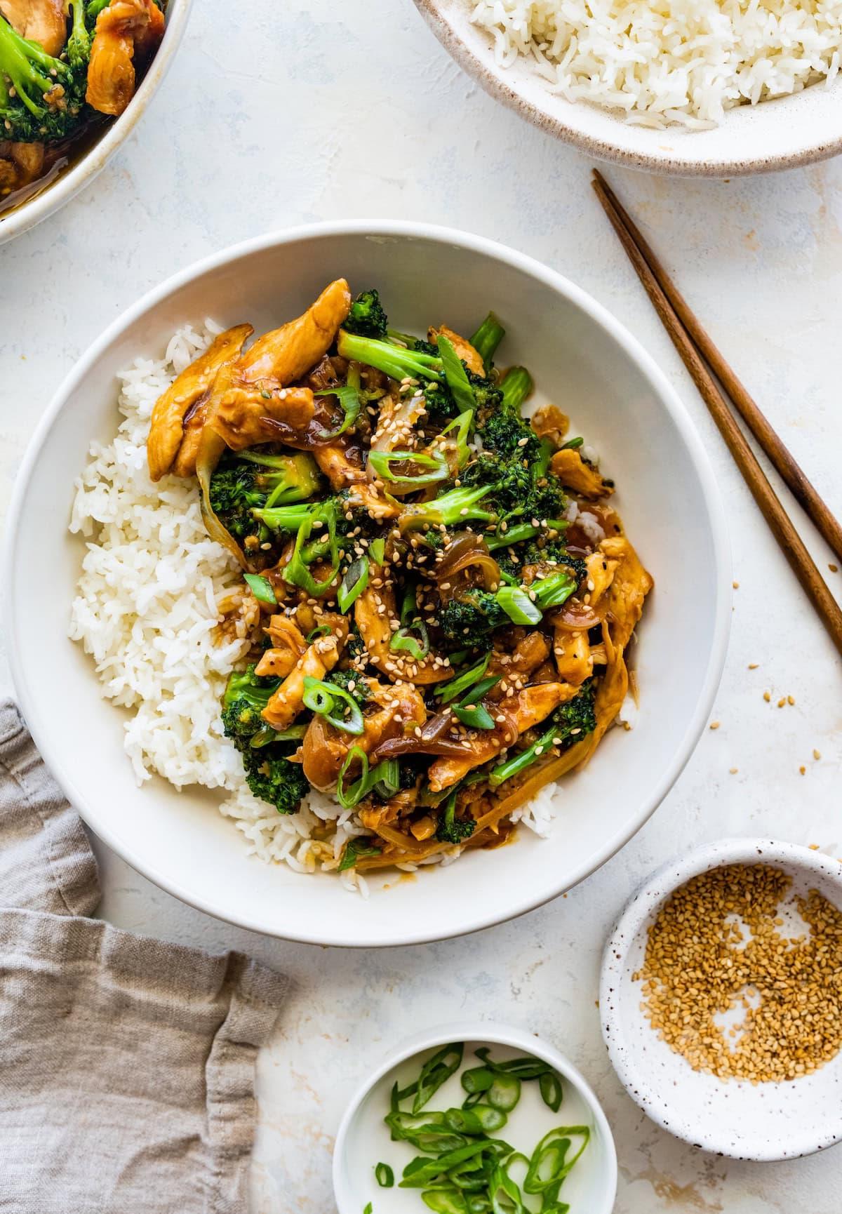 **Κοτόπουλο και Μπρόκολο Σοτέ** 8 Κοτόπουλο stir fry με μπρόκολο σε λευκό μπολ, σερβιρισμένο με λευκό ρύζι και γαρνιρισμένο με σουσάμι και φρέσκο κρεμμυδάκι. Ξύλινα chopsticks είναι δίπλα στο μπολ.