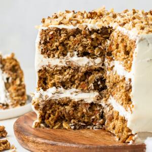 Κέικ βρώμης και καρότου 9 3-layer oatmeal carrot cake, frosted with chopped walnuts on top served on a wooden board. A few slices of the cake have been cut out exposing the inside of the cake.