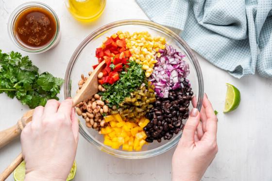 Ένα χέρι ανακατεύει τα υλικά της σαλάτας cowboy caviar σε ένα μεγάλο γυάλινο μπολ.