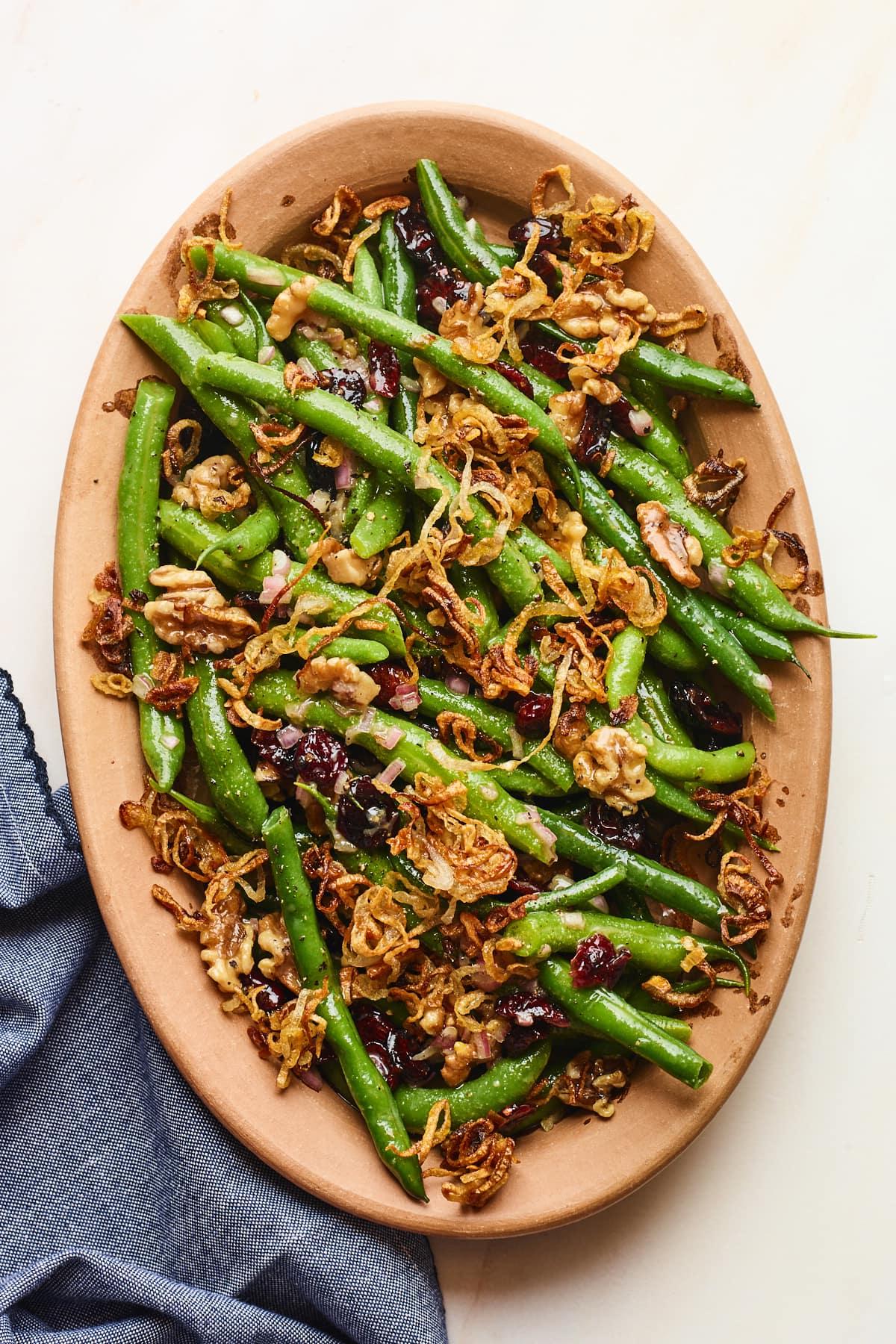 Φασολάκια Σαλάτα 10 An overhead photo looking down onto a serving dish containing green bean salad topped with fried shallots. A blue linen dish cloth is laying next to the serving dish.