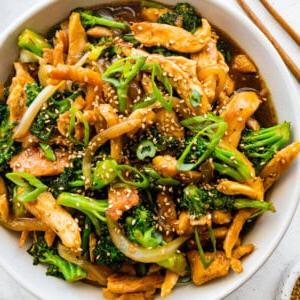 **Κοτόπουλο και Μπρόκολο Σοτέ** 10 Κοτόπουλο stir fry με μπρόκολο σε λευκό μπολ, γαρνιρισμένο με σουσάμι και φρέσκο κρεμμυδάκι, με ξύλινα chopsticks δίπλα.