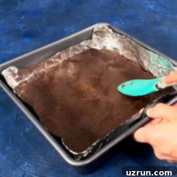 Five-Layer Chocolate Cookie Bars 5 Chocolate cookie dough being spread in square baking pan with spatula.