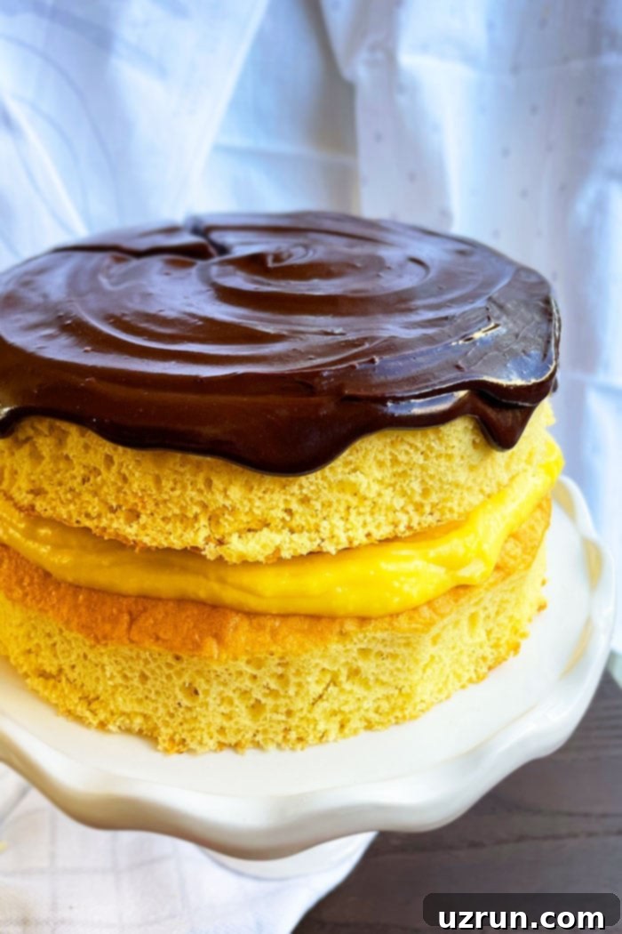 Easy homemade Boston Cream Pie on a white cake stand, showcasing its layers of yellow cake, vanilla pudding, and chocolate ganache