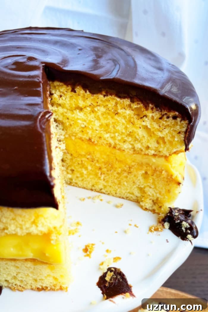 Easy Boston Cream Pie 3 Closeup shot of a Boston Cream Pie with a slice removed, revealing the perfect layers of yellow cake and vanilla cream, on a white cake stand