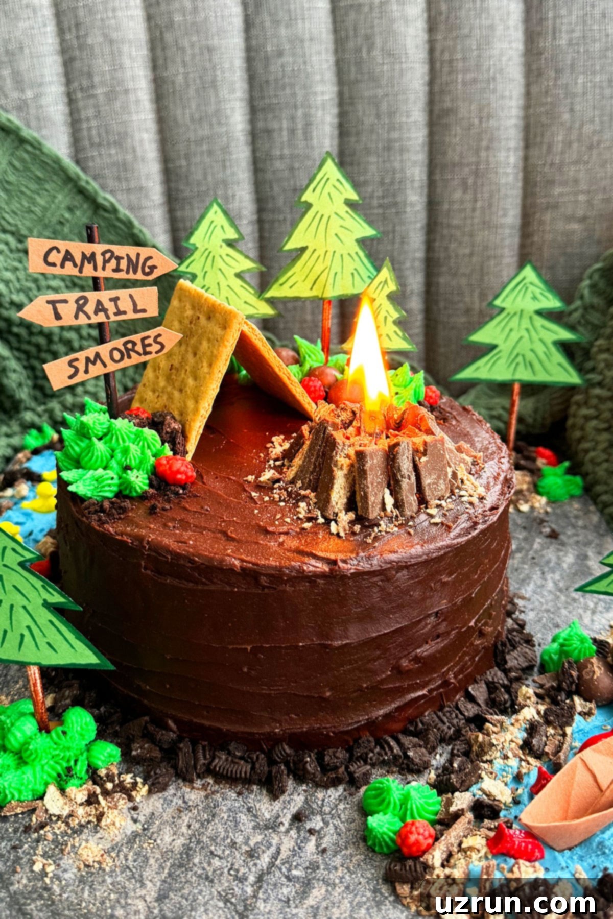 Easy Camping Cake featuring a realistic, edible tent and a lit miniature campfire on a rustic gray cake board, surrounded by candy trees and crushed cookie 'dirt'.